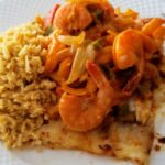 A-grilled-fish-fillet-topped-with-sautéed-shrimp,-onions,-and-bell-peppers,-served-alongside-a-portion-of-seasoned-rice-on-a-white-plate.