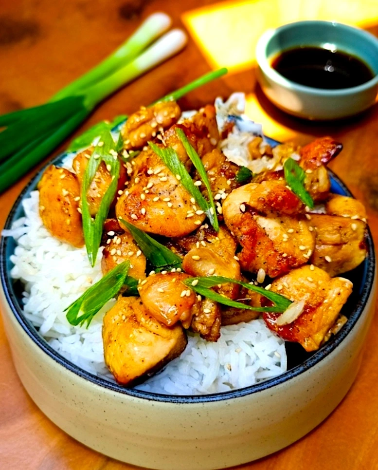 A-bowl-of-fluffy-white-rice-topped-with-glistening-golden-brown-sticky-soy ginger chicken-pieces,-garnished-with-fresh-green-onions-and-sesame-seeds.-A-small-bowl-of-extra-sauce-is-visible-in-the-background.