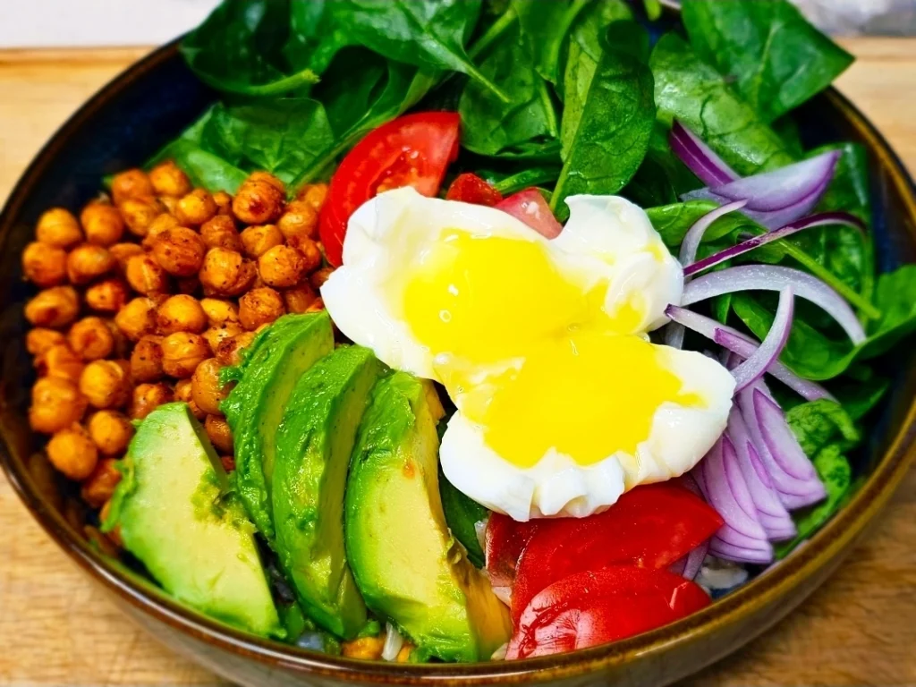A-healthy-veggie power bowl-filled-with-roasted-chickpeas,-vibrant-spinach,-sliced-avocado,-red-onions,-and-a-poached-egg-with-a-runny-golden-yolk.